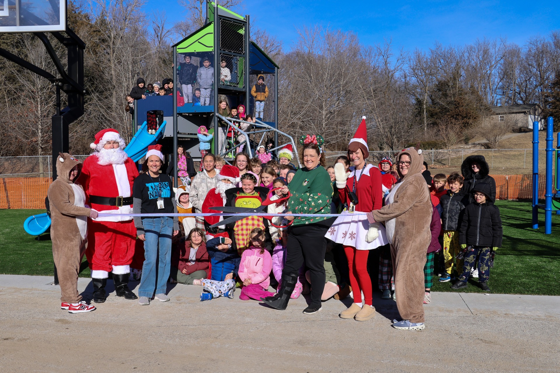 A group of students and adults posing in front of the new playground equipment. Two adults are holding a blue ribbon spread across the front and another adult holding large scissors.
